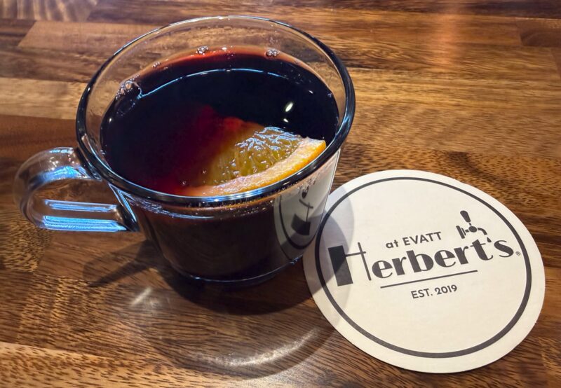 A glass mug of mulled wine with an orange slice, beside a Herbert's at Evatt coaster on a wooden table.